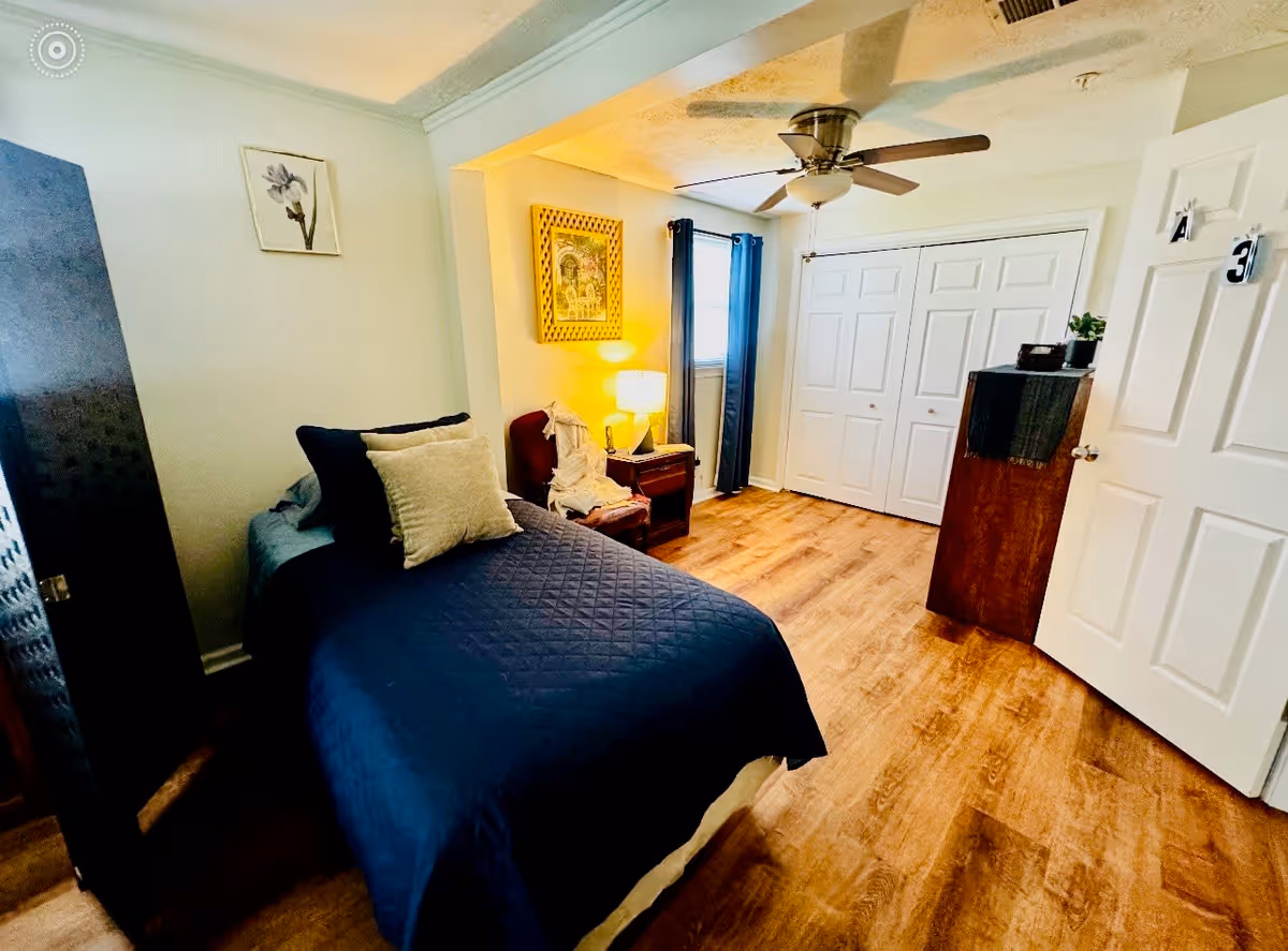 Small furnished bedroom with a single bed, nightstand and lamp, ceiling fan, closet doors and wood-look flooring.