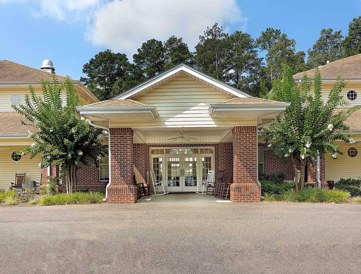 Front exterior view of a memory care community building with a covered entrance supported by brick columns, rocking chairs on the porch, and trees on either side under a partly cloudy sky.