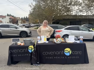 A woman stands behind two black tables covered with black tablecloths displaying the Covenant Care Home Health & Hospice logo. The tables have informational pamphlets, a basket, and other promotional materials. The setting is an outdoor parking lot with several parked cars and trees in the background.