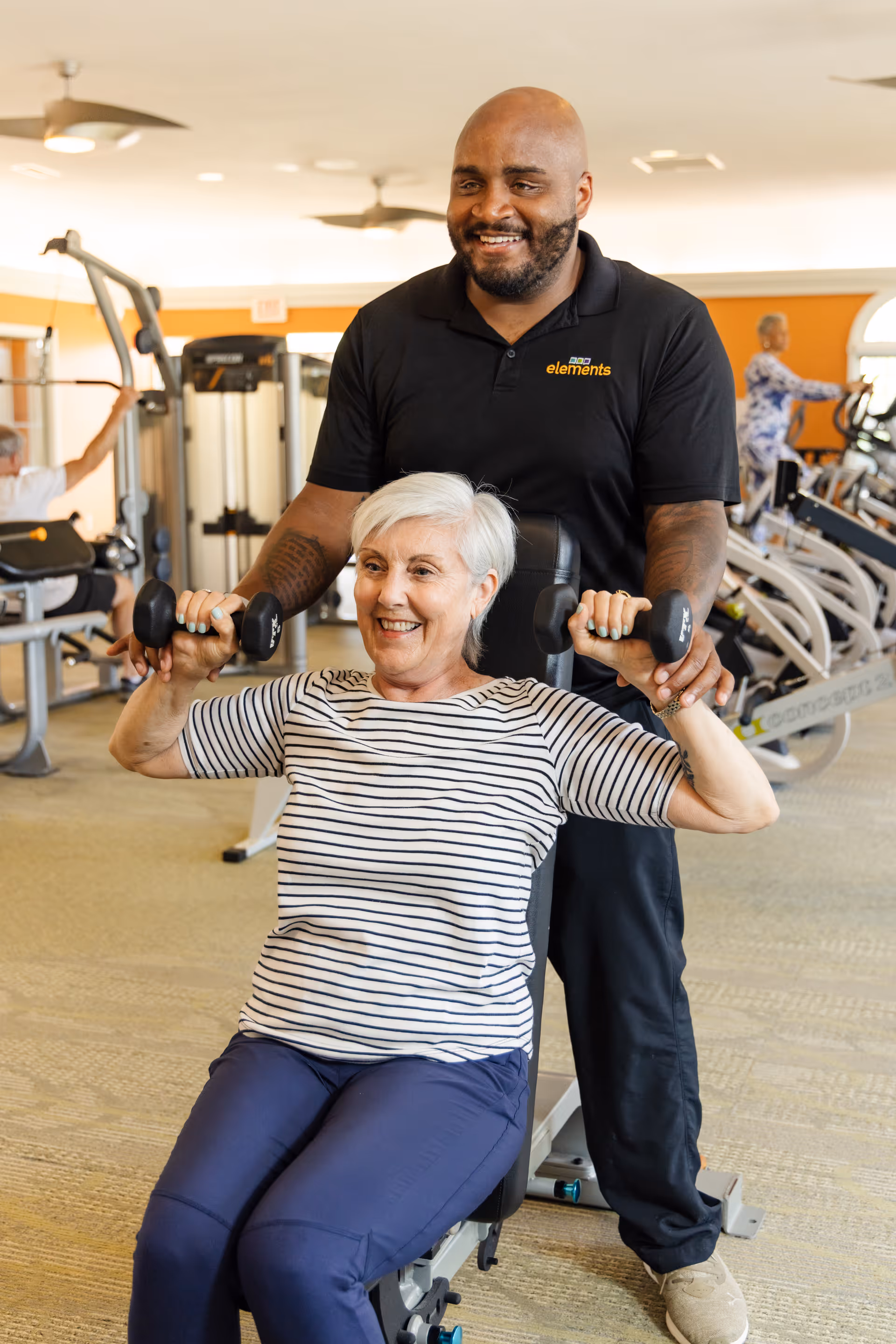 An elderly woman in a striped shirt and blue pants is seated on a workout bench lifting dumbbells with the assistance of a smiling male trainer wearing a black polo shirt with the logo 'elements'. They are in a well-lit gym with exercise equipment and other people working out in the background.