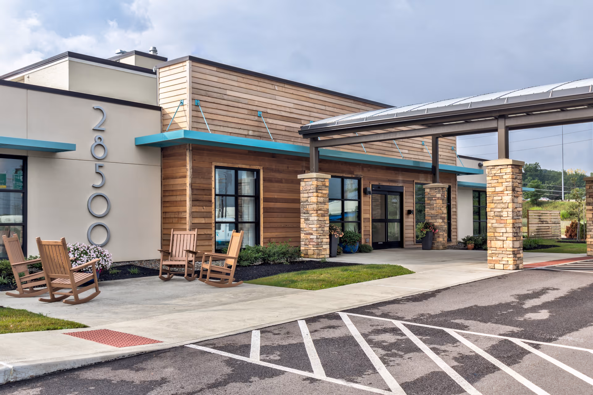 Exterior view of a modern senior living facility entrance with stone pillars, wooden siding, large windows, and a covered drop-off area. There are wooden rocking chairs and potted plants near the entrance, with a parking area in the foreground.