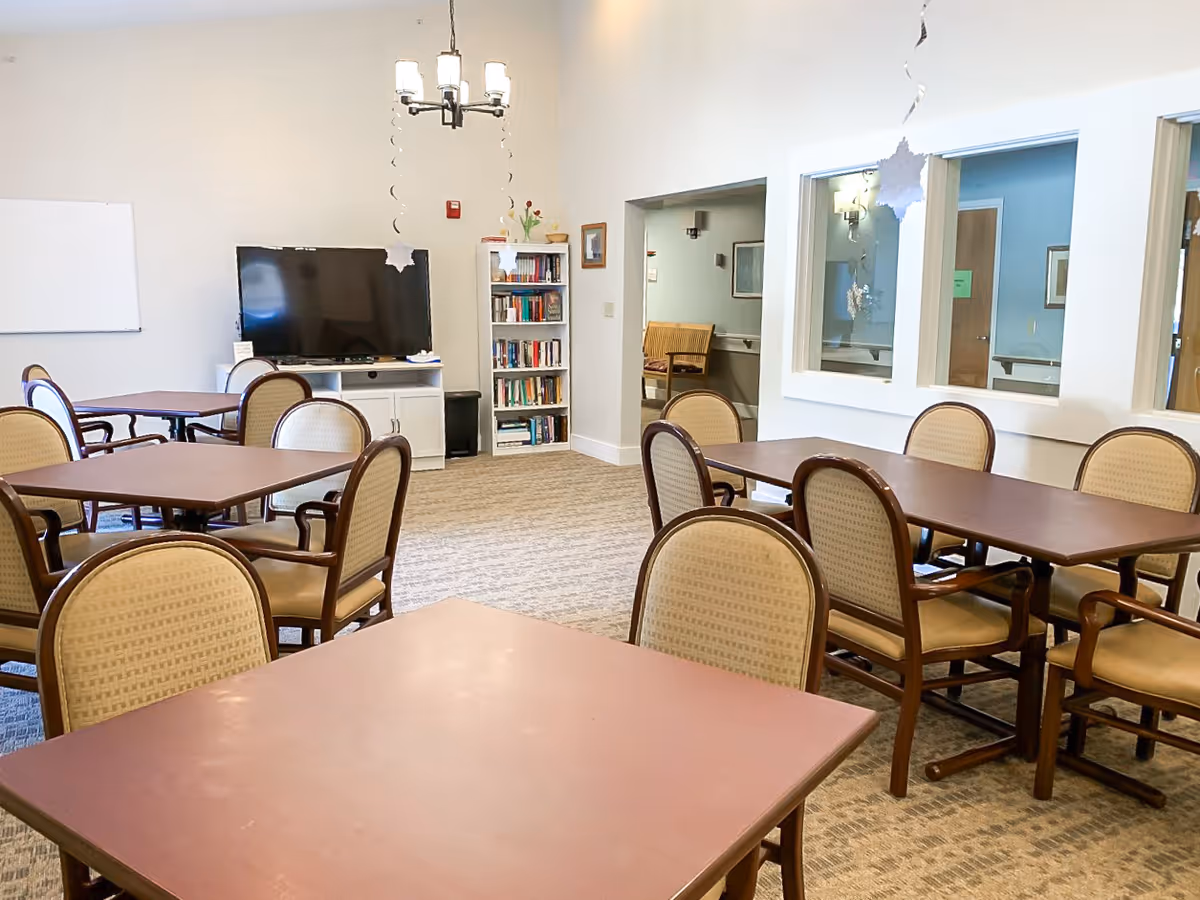 A common area in The Lodges Care Center featuring multiple tables with chairs arranged around them. There is a large flat-screen TV on a white stand, a bookshelf filled with books, and a chandelier hanging from the ceiling. The room has beige walls and carpeted flooring, with windows looking into adjacent rooms.