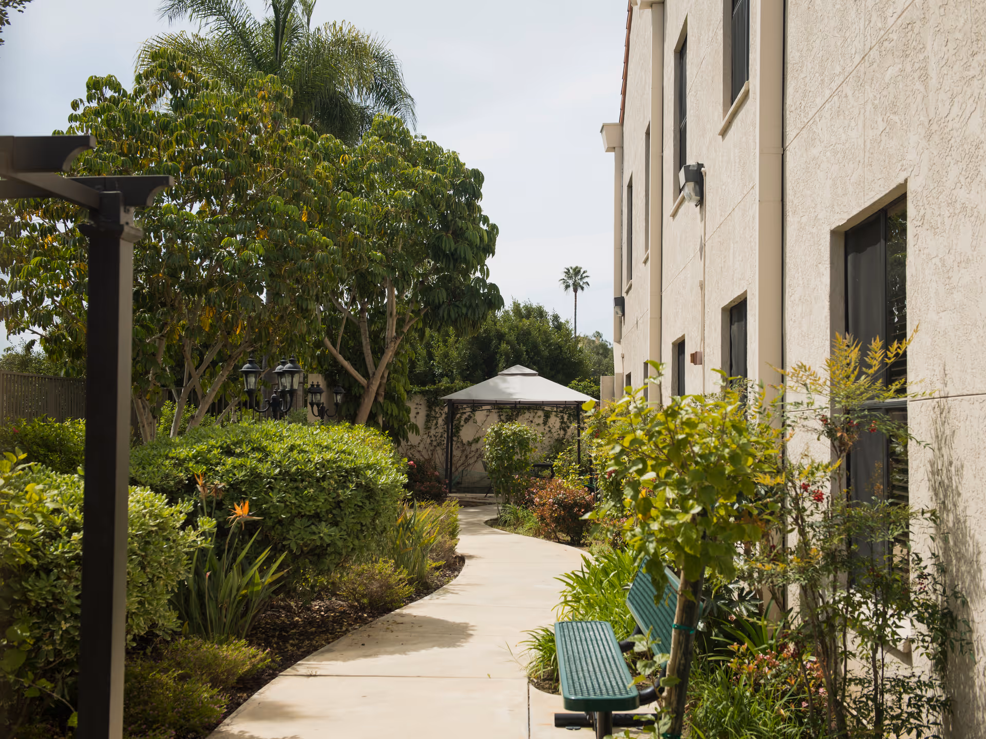 A winding concrete pathway through a garden area beside a beige building. The path is lined with green bushes, trees, and plants. There is a green bench on the right side and a gazebo with a white canopy in the background. The sky is clear and the scene is well-lit with natural daylight.