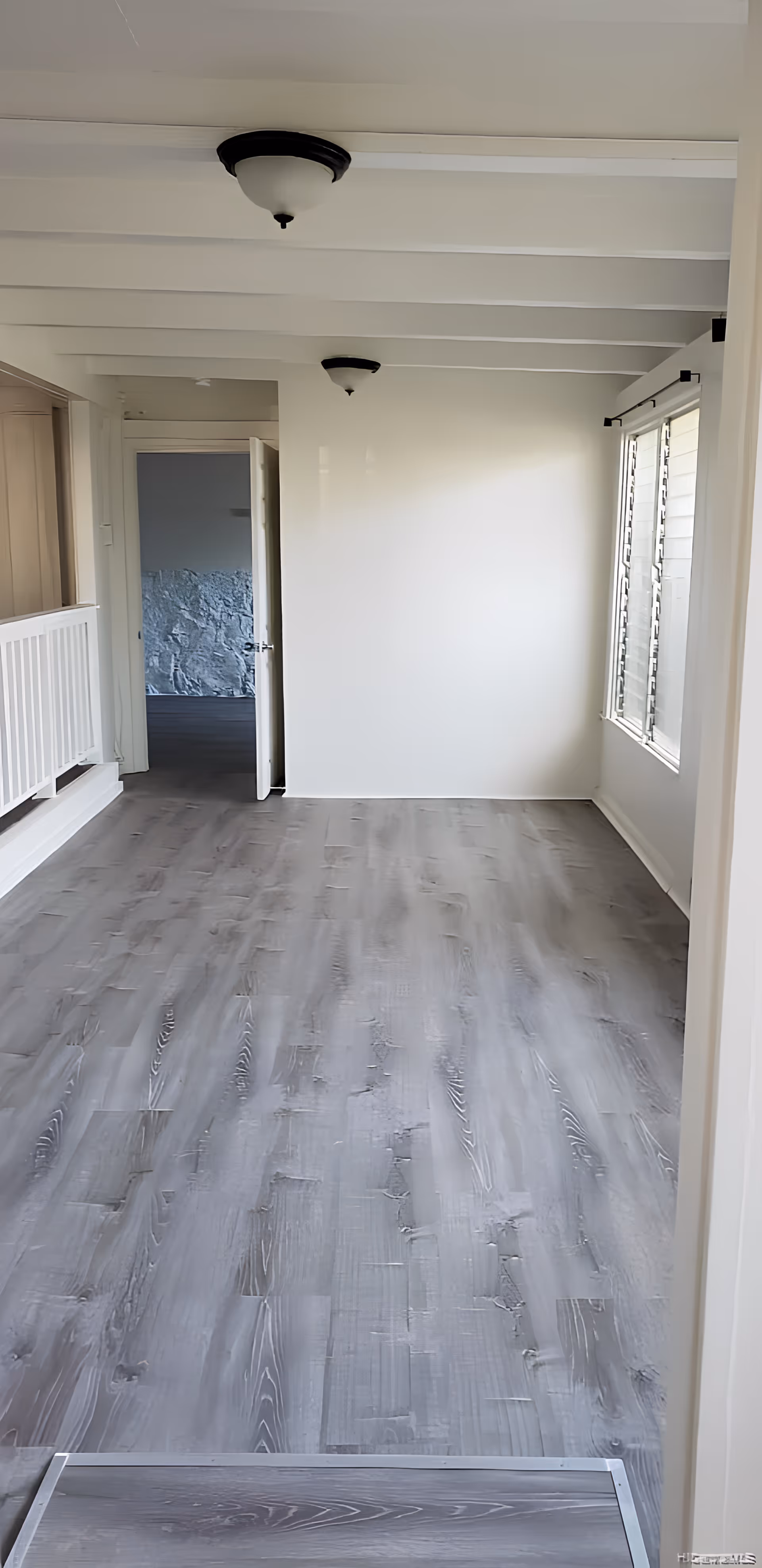 Empty interior room with gray wooden flooring, white walls, and ceiling beams. Two ceiling light fixtures are visible. There is a white railing on the left side and a window with blinds on the right side. An open door leads to another room with a textured wall.