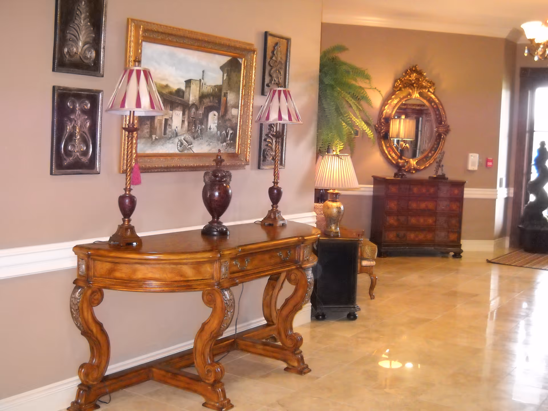 A richly furnished lobby with an ornate wooden console table, lamps, framed artwork, a decorative chest and mirror on a polished tile floor.