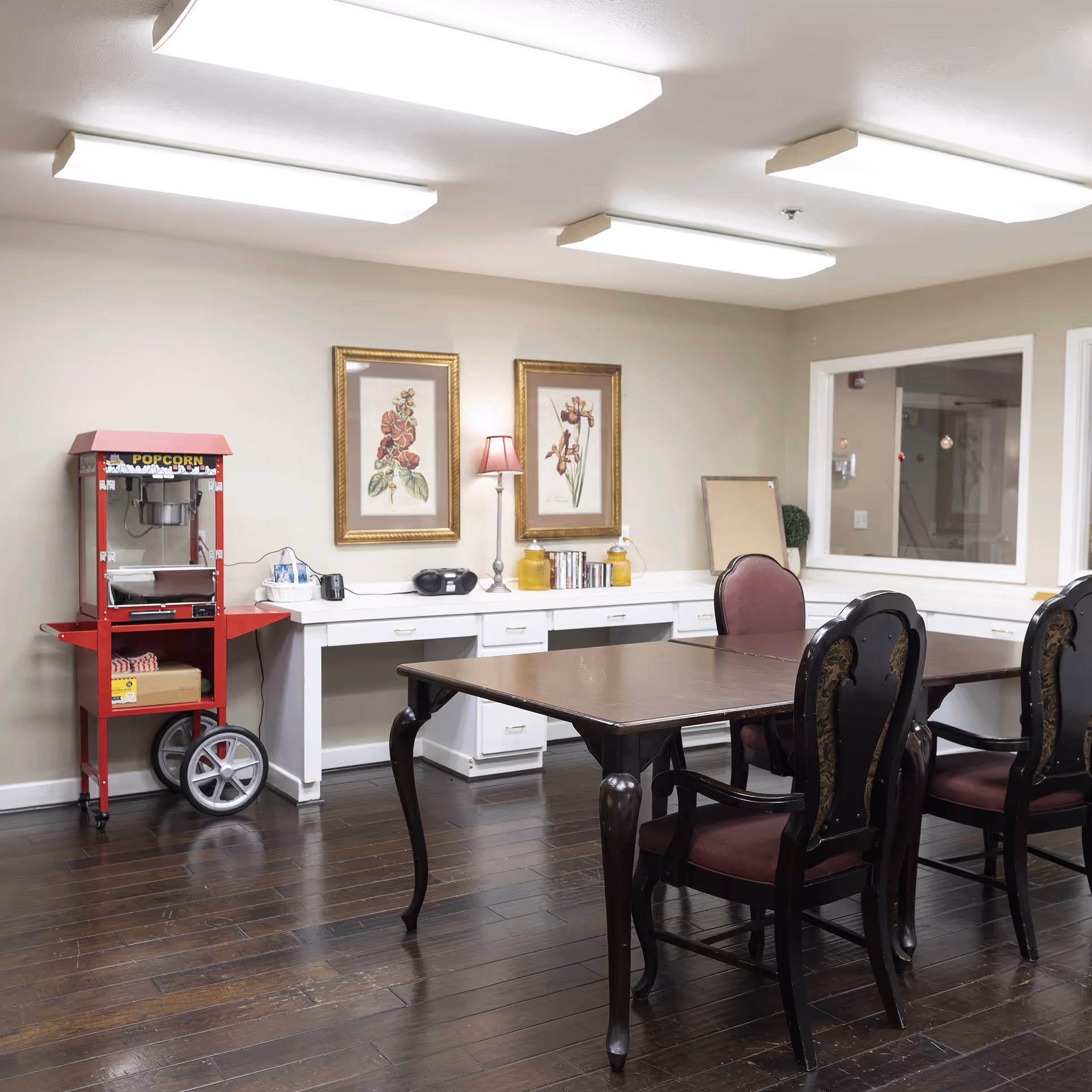 A dining/activity room with a wooden table and upholstered chairs, a red popcorn machine, framed floral prints, and white counter space under fluorescent lights.