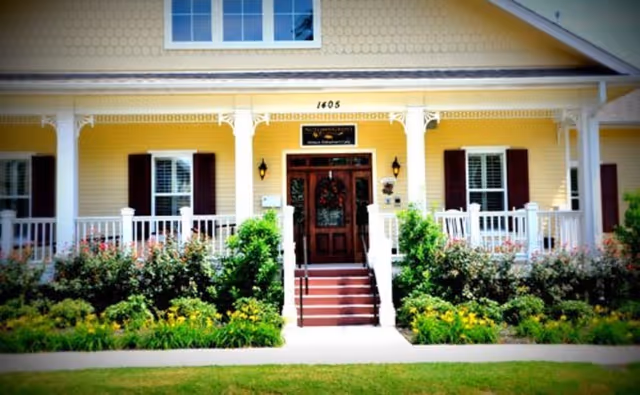 Front exterior view of a yellow building with white trim, a porch with white railings, steps leading to a dark wooden door, windows with dark shutters, and well-maintained bushes and flowers in front.