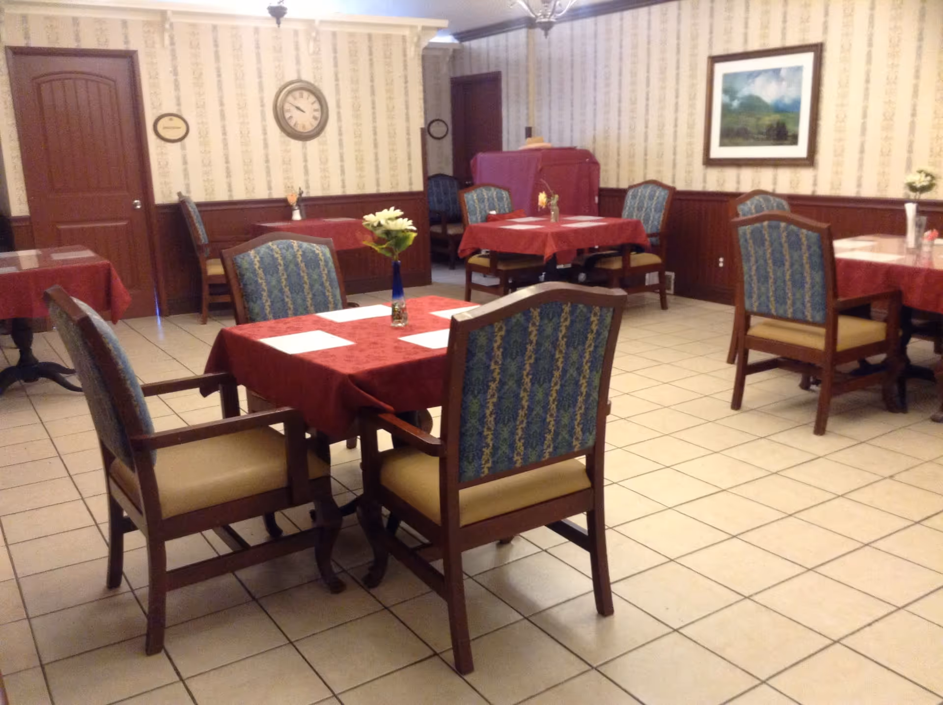 A dining room with several tables covered in red tablecloths, each set with placemats and a small vase with flowers. The room has tiled floors, wallpapered walls with a clock and framed painting, and wooden chairs with blue patterned upholstery.