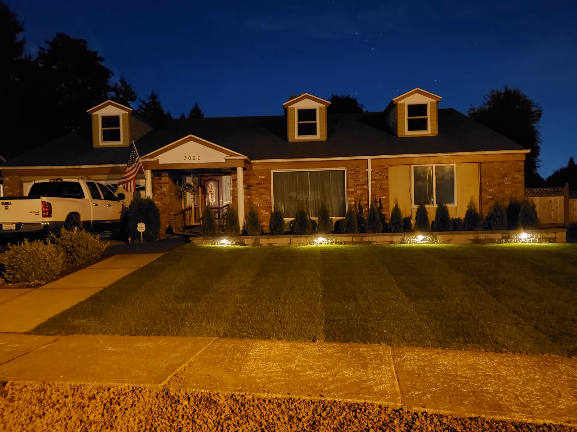 Nighttime view of a single-story brick house with three dormer windows on the roof, a white front door with decorative glass, an American flag near the entrance, a white pickup truck parked in the driveway, a well-maintained lawn with small shrubs and outdoor lighting along the front, and a sidewalk in the foreground.
