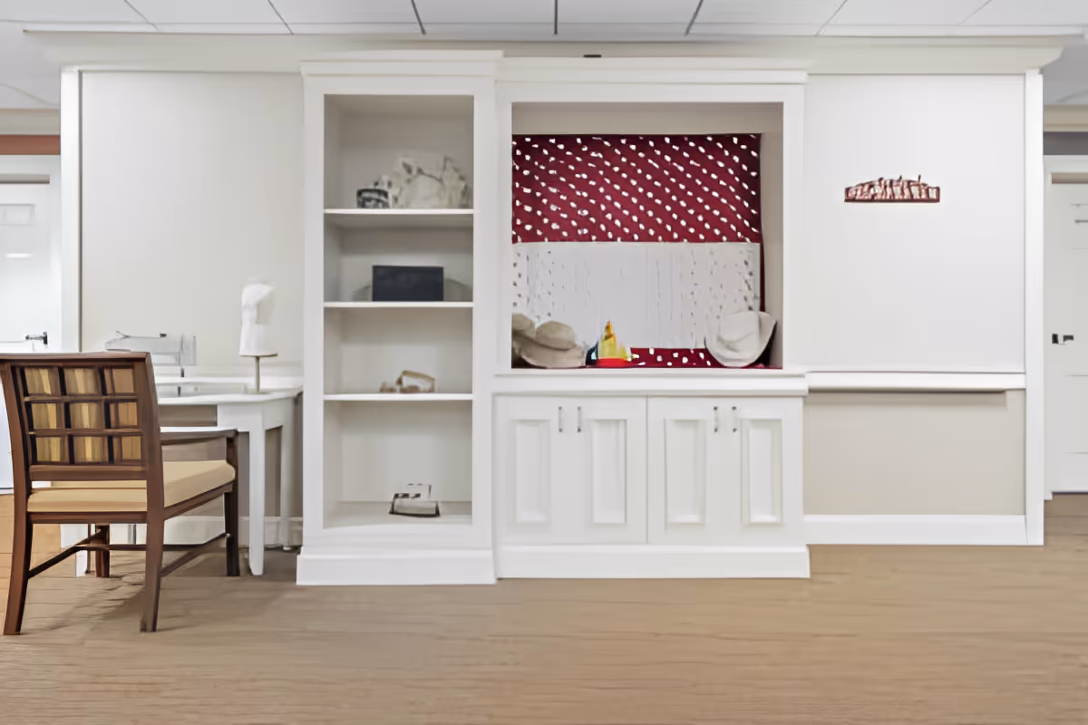 A room with white built-in shelves and cabinets, a wooden chair with a cushioned seat, and a small white table. The shelves contain a few decorative items, and there is a red and white patterned fabric or panel in the middle section of the built-in unit. The floor is carpeted in a light brown color.