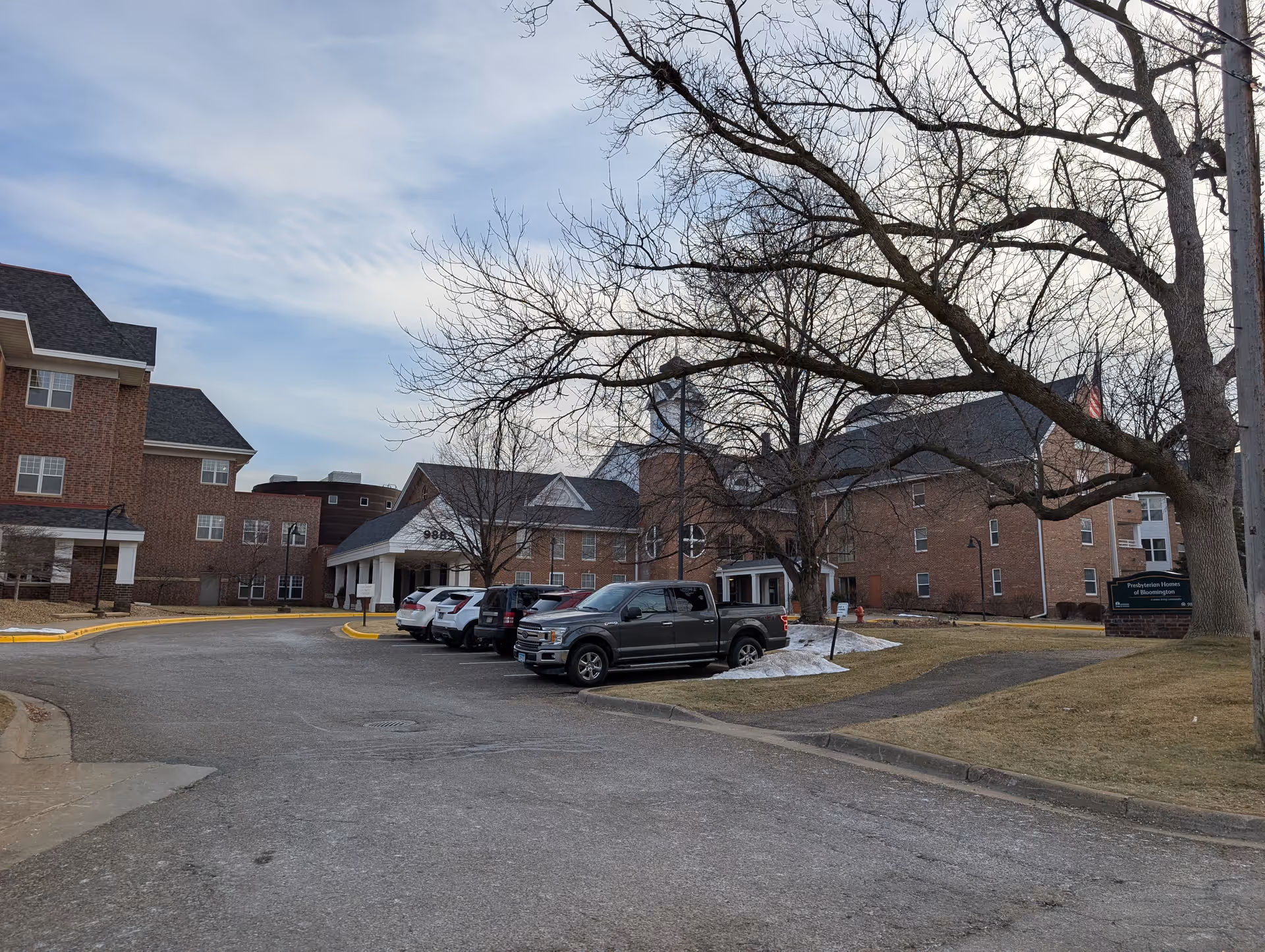 Brick senior living building with a covered entrance, parked cars, and leafless trees under a cloudy sky.