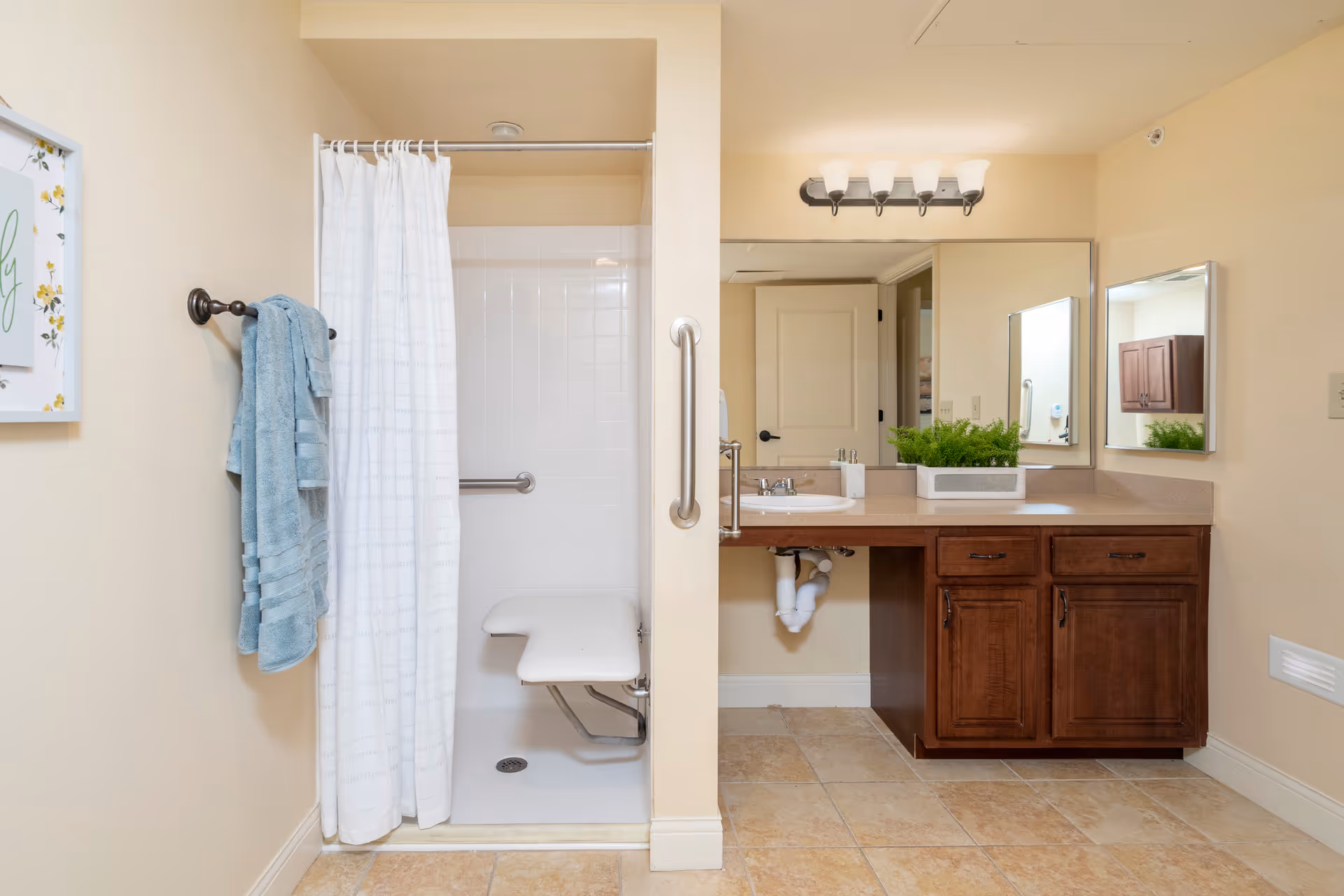 A bathroom with a walk-in shower featuring a white shower curtain and a fold-down seat. There is a towel rack on the left wall with a blue towel hanging. On the right side, there is a wooden vanity with a sink, a large mirror above it, and a small green plant on the countertop. The floor is tiled in a beige color.