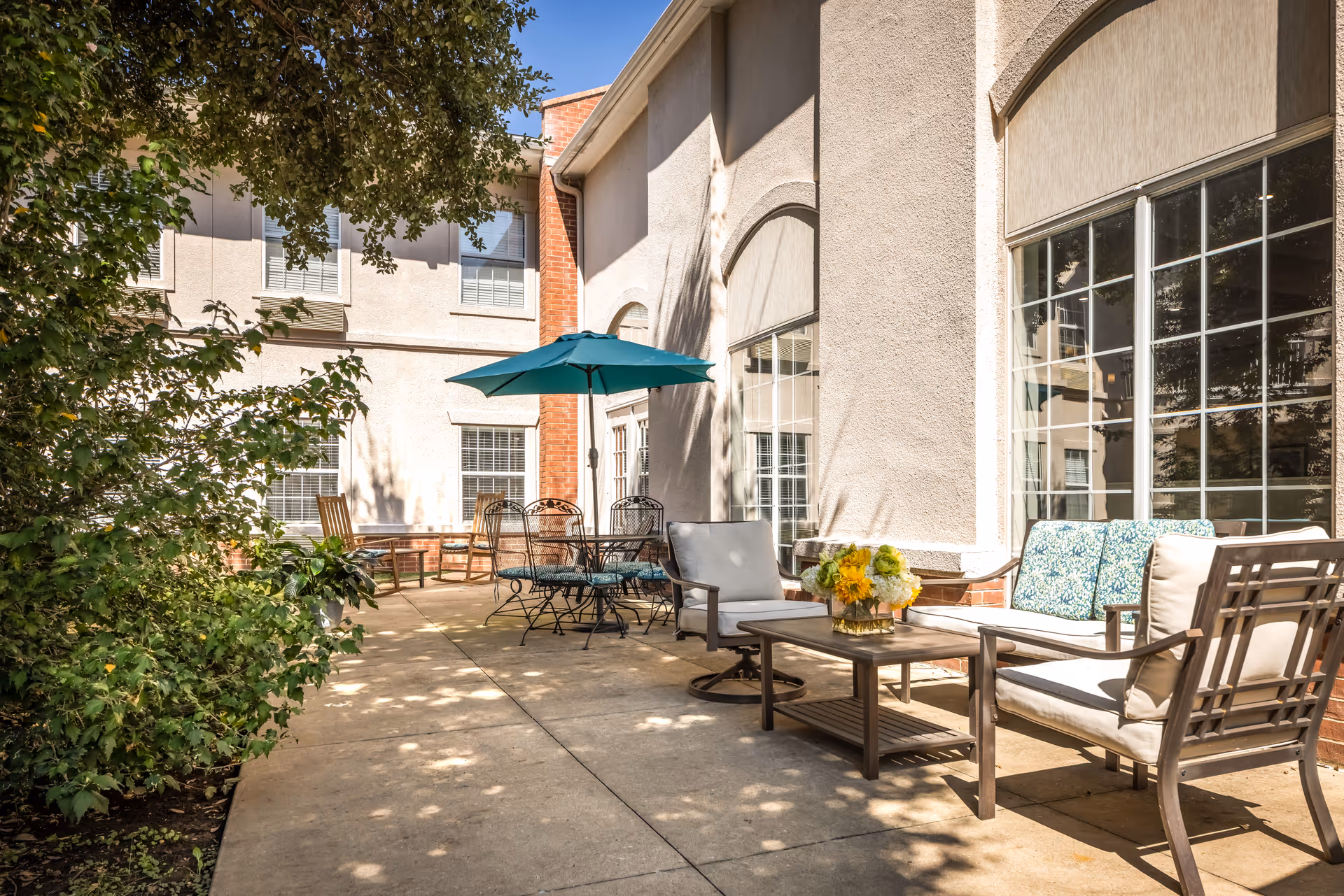Outdoor patio area at The Waterford at Plano with cushioned seating, a coffee table with a flower arrangement, a round table with metal chairs, and a teal umbrella. The patio is adjacent to a building with large windows and surrounded by greenery.