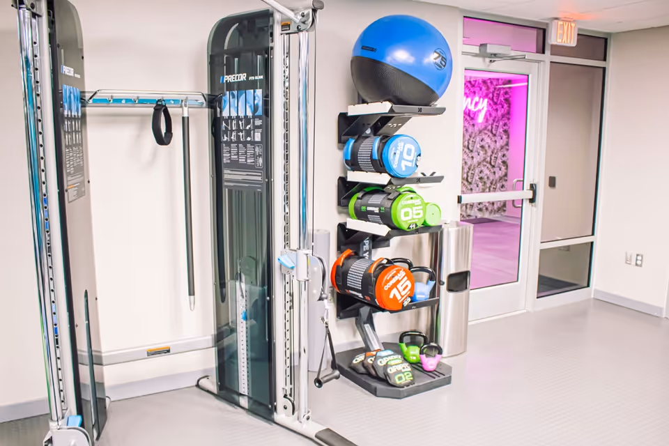 Interior view of a fitness room with exercise equipment including a Precor cable machine, a rack holding various weighted exercise balls, kettlebells, and other fitness accessories. A glass door with an exit sign above it leads to another room illuminated with pink lighting.