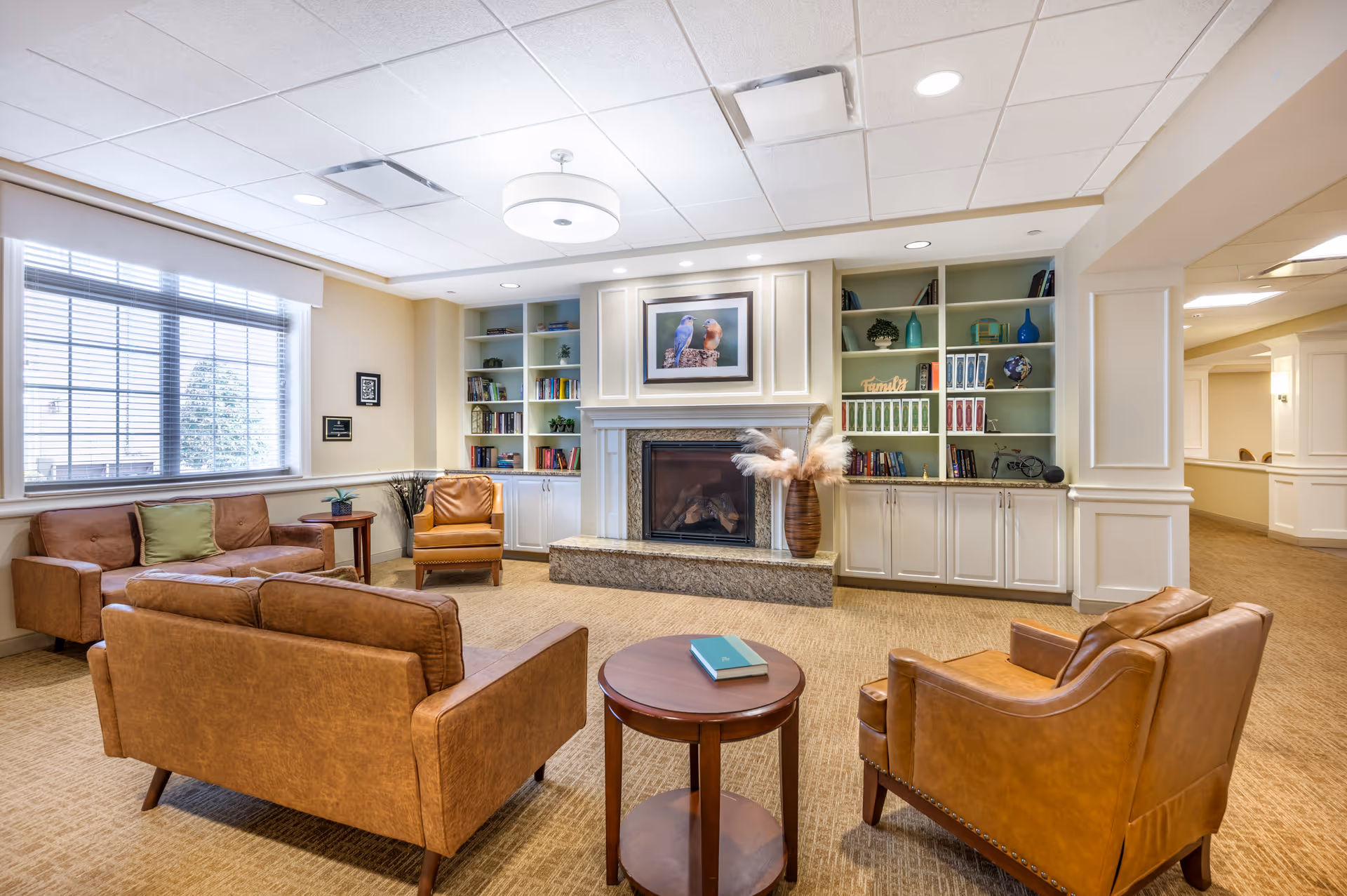 A cozy living room area in a senior living facility with tan leather sofas and armchairs arranged around a small wooden round table. The room features a fireplace with a framed picture of two birds above it, built-in white cabinets and shelves filled with books and decorative items, and a large window with blinds letting in natural light.