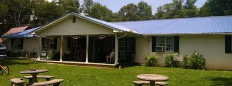 Single-story building with a covered front porch and circular picnic tables on a green lawn.