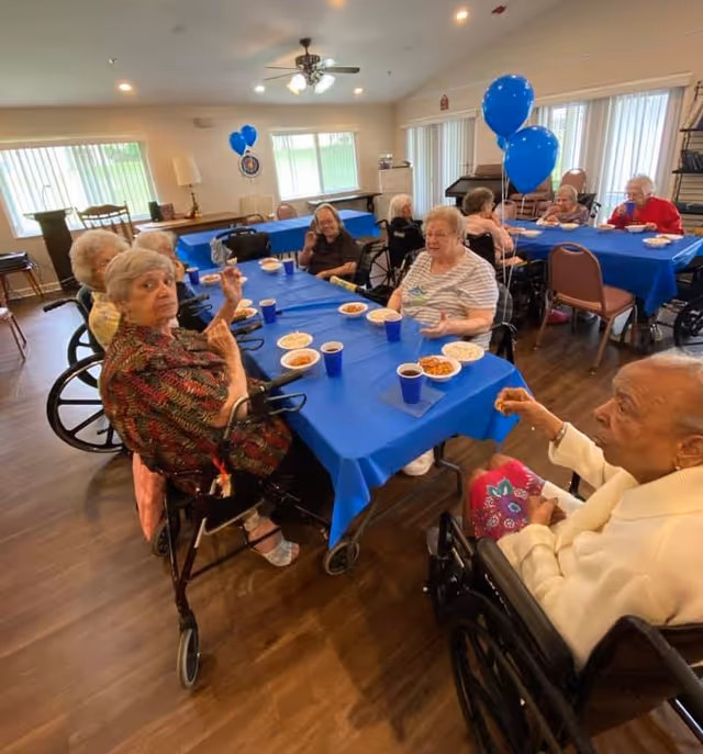 A group of elderly people sitting around tables covered with blue tablecloths in a well-lit room, enjoying a meal with plates of food and blue cups. Some individuals are in wheelchairs, and there are blue balloons tied to chairs in the background.