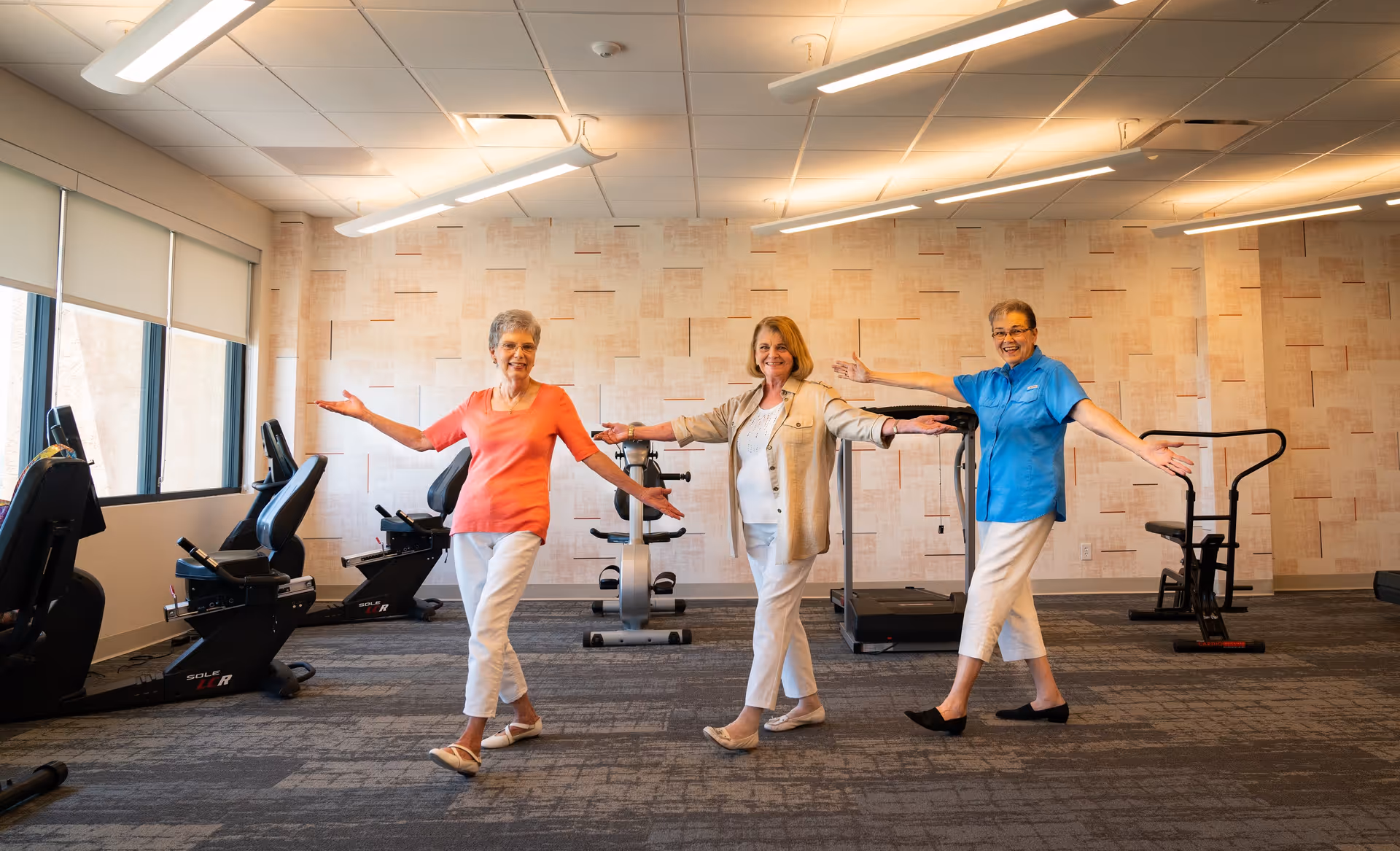 Three elderly women standing in a fitness room with exercise equipment, smiling and posing with arms outstretched. The room has large windows with blinds, a patterned wall, and overhead fluorescent lighting.