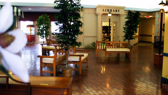 Indoor common area with wooden benches, potted trees, and a doorway labeled 'LIBRARY'.