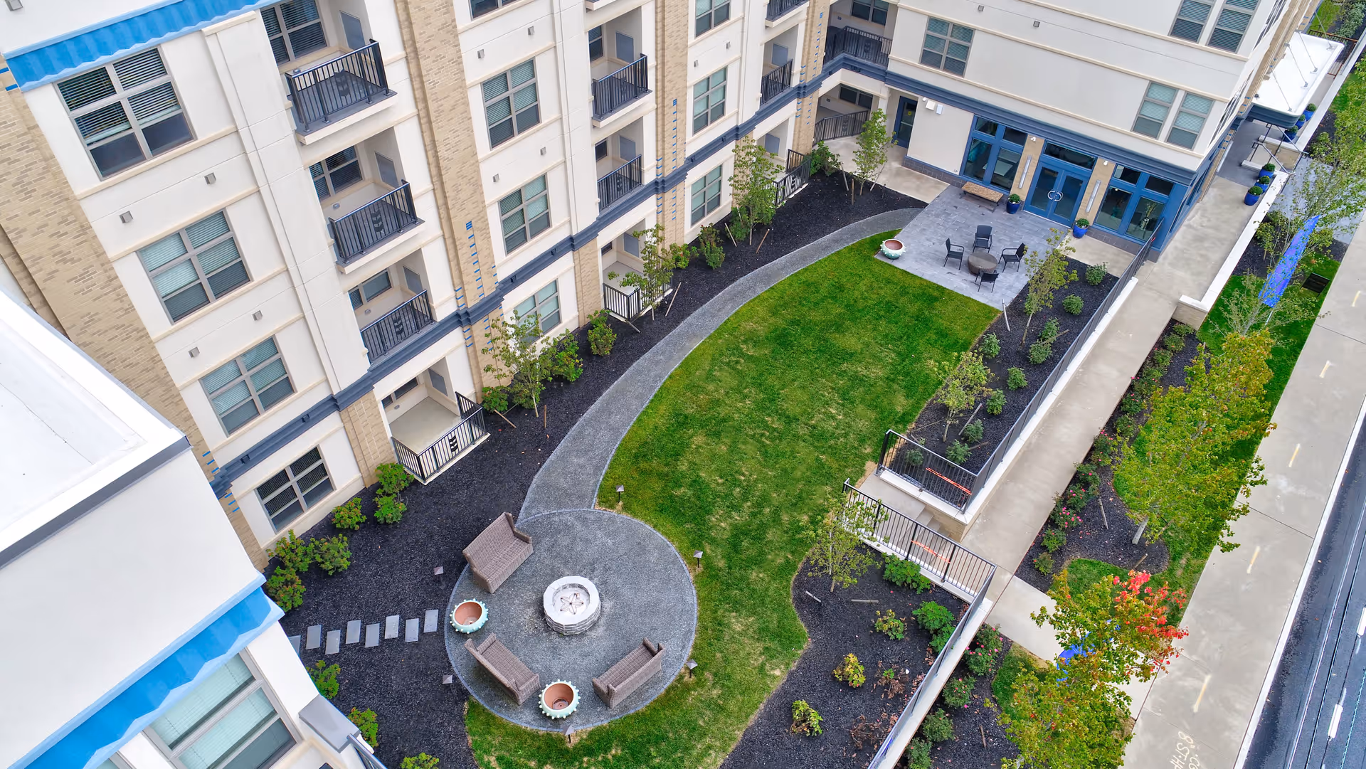 Aerial view of a landscaped courtyard between multi-story residential buildings with a circular seating/fire pit area, lawn, pathways, and patio seating.
