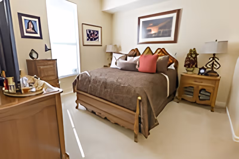 A cozy bedroom with a wooden bed frame covered in a brown bedspread and multiple pillows, including a red one. There are two wooden nightstands on either side of the bed, each with a lamp and decorative items. A dresser with various items on top is positioned near a window with white blinds. The walls are adorned with framed artwork.