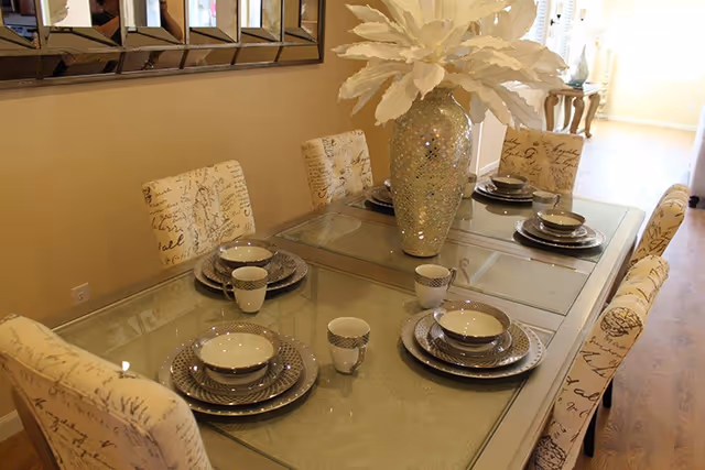 A dining table set for six with matching plates, bowls, and cups. The table has a glass top and a large decorative vase with white leaves as a centerpiece. The chairs around the table are upholstered with fabric featuring script writing. A large mirror hangs on the wall behind the table.