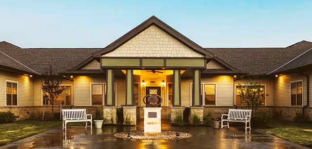 Front exterior view of Silverado Overland Park Memory Care Community building at dusk with warm lights illuminating the entrance. The building features a peaked roof, stone and siding facade, two white benches on either side of the entrance, potted plants, and a small statue on a pedestal in the center of the walkway.