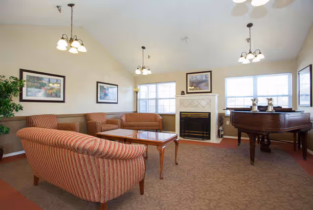 A spacious living room with beige walls and carpeted floor featuring a striped sofa and loveseat, a wooden coffee table, a fireplace with a framed picture above it, a grand piano, and several windows allowing natural light to fill the room.