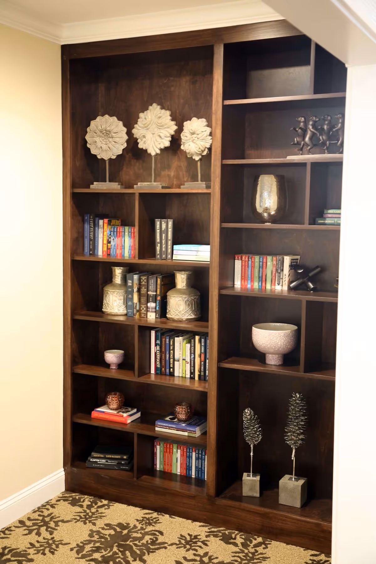 A dark wooden bookshelf filled with various books and decorative items including sculptures, vases, and small art pieces, set against a beige wall with a patterned carpet on the floor.