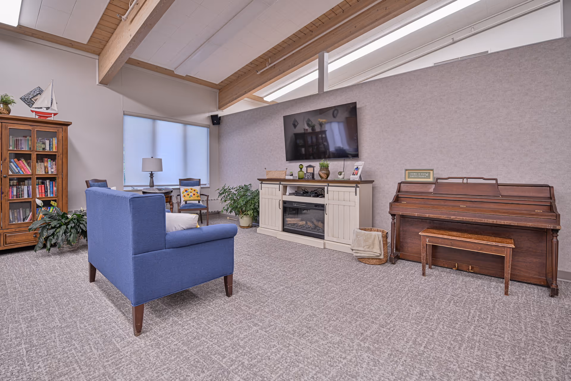 A cozy living room area with a blue armchair facing a wall-mounted flat-screen TV above a white cabinet with decorative items. To the right, there is a wooden piano with a matching bench and a small framed sign on top. On the left side, there is a wooden bookshelf filled with books and a model sailboat on top, next to a window with a lamp and two chairs. The room has a carpeted floor, light-colored walls, and a ceiling with wooden beams and skylights.