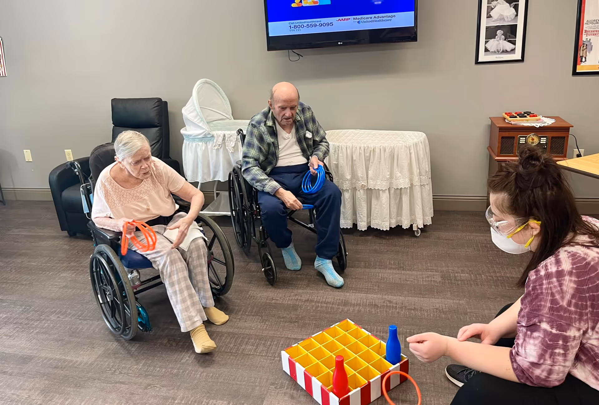 Two elderly individuals in wheelchairs playing a ring toss game with a caregiver wearing a face mask and protective eyewear in a senior care facility room with a TV mounted on the wall and a bassinet in the background.