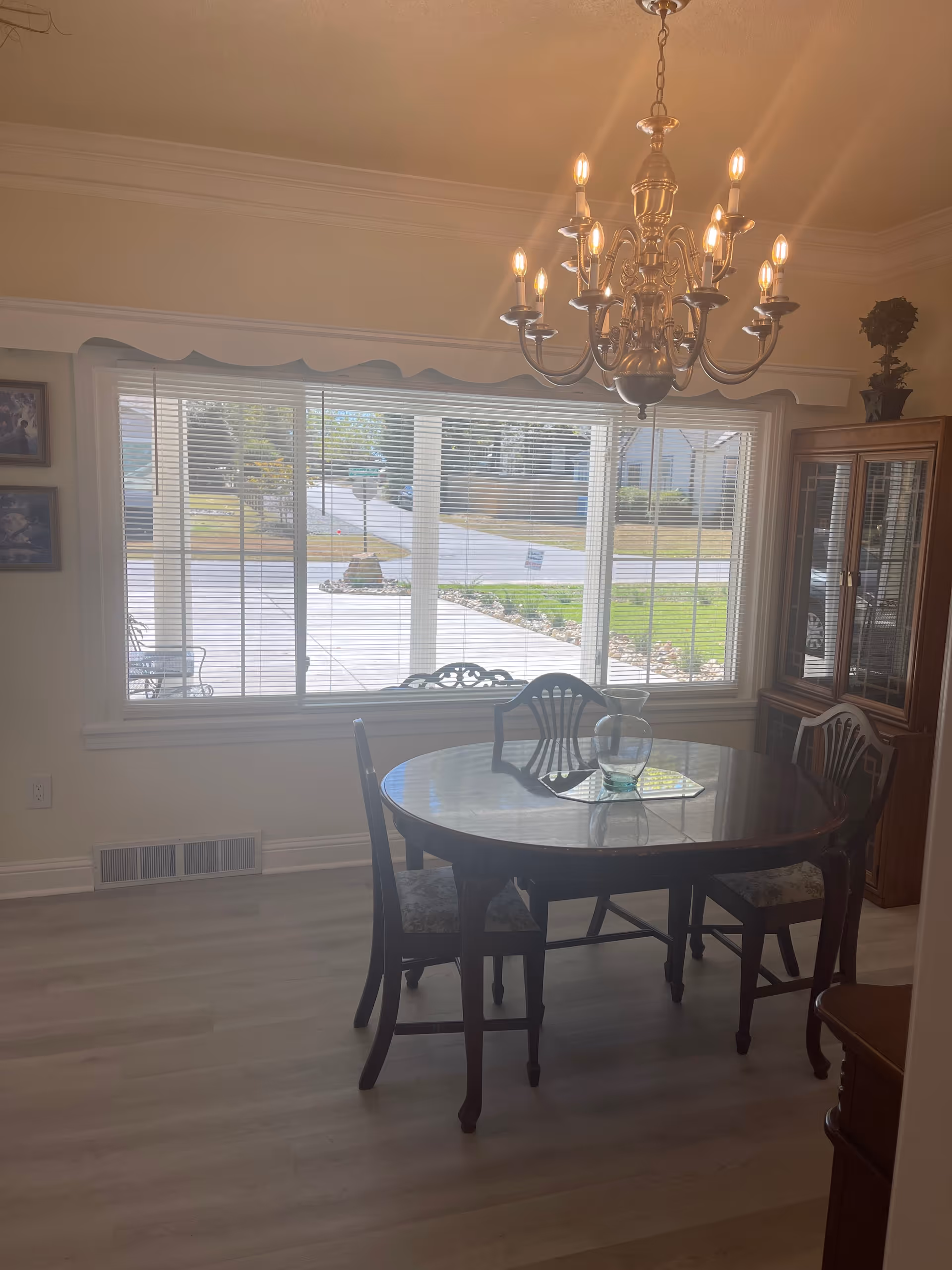 A dining room with a round wooden table and four chairs. A glass vase is placed on the table. A large window with blinds lets in natural light, showing a view of a driveway and lawn outside. A chandelier with multiple lit bulbs hangs from the ceiling. A wooden cabinet with glass doors is visible in the corner.