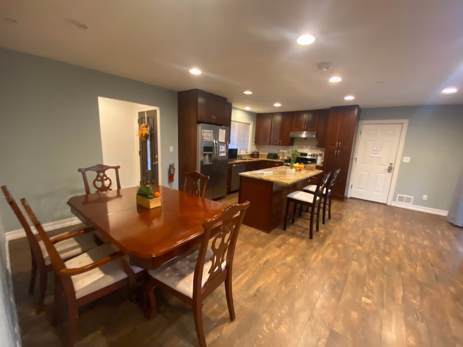 Interior view of a senior living facility kitchen and dining area featuring a wooden dining table with six chairs, a kitchen island with three stools, dark wood cabinets, stainless steel appliances including a refrigerator and stove, and wood flooring.