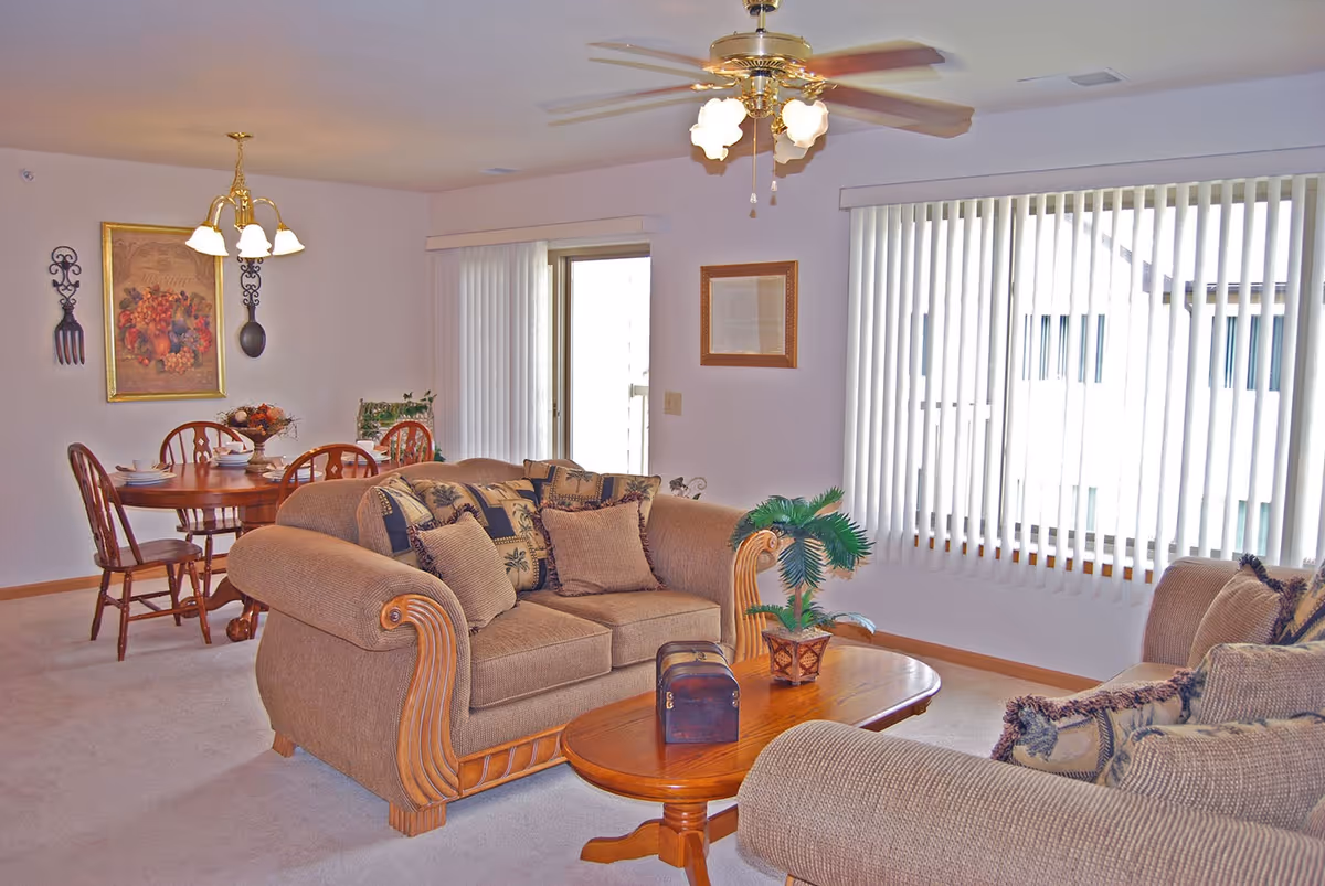 Open-plan living room and dining area with beige sofas, a wooden coffee table, dining set, and vertical blinds on the windows.