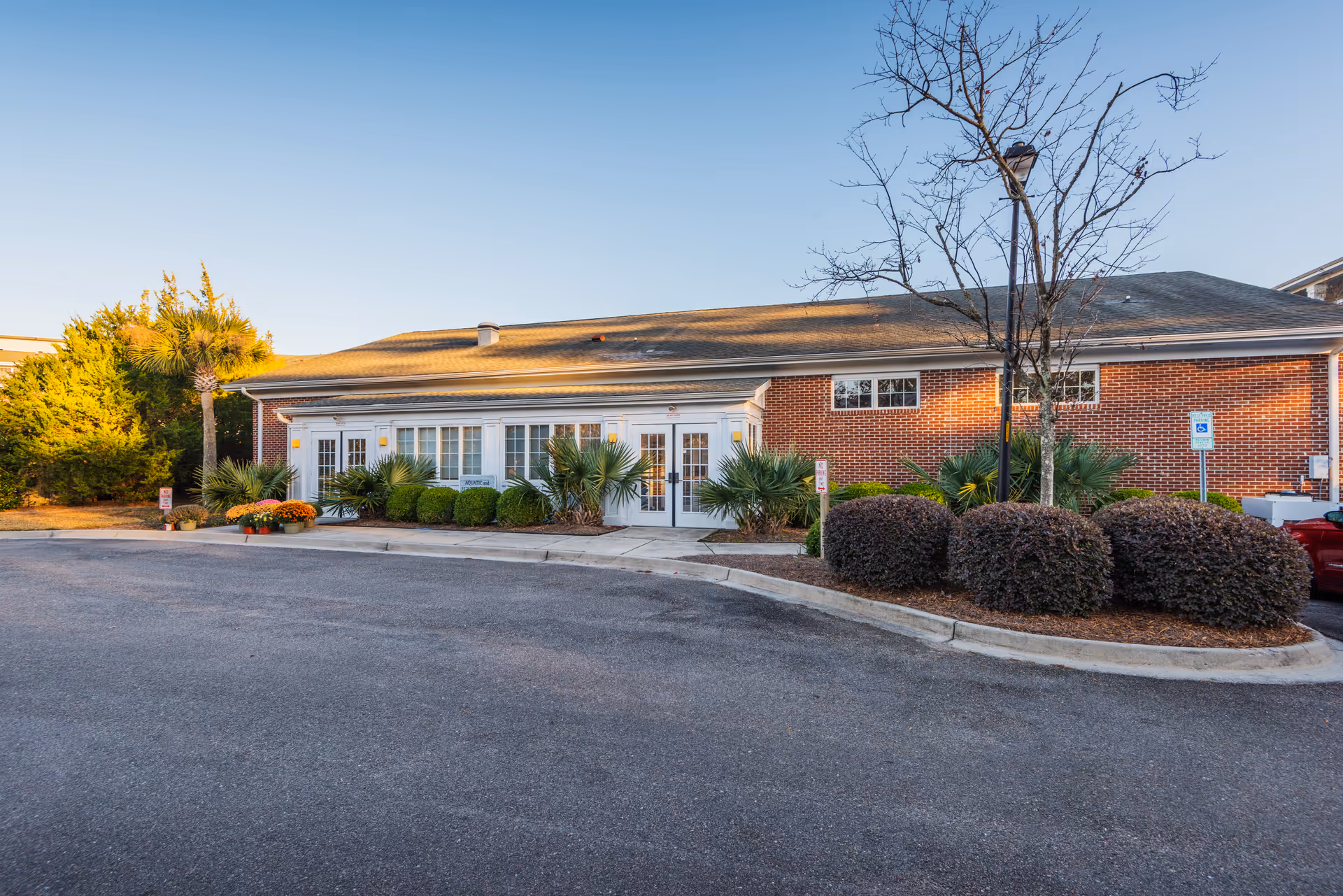 Front exterior of a single-story brick senior living building with glass doors, landscaped shrubs and a parking area.