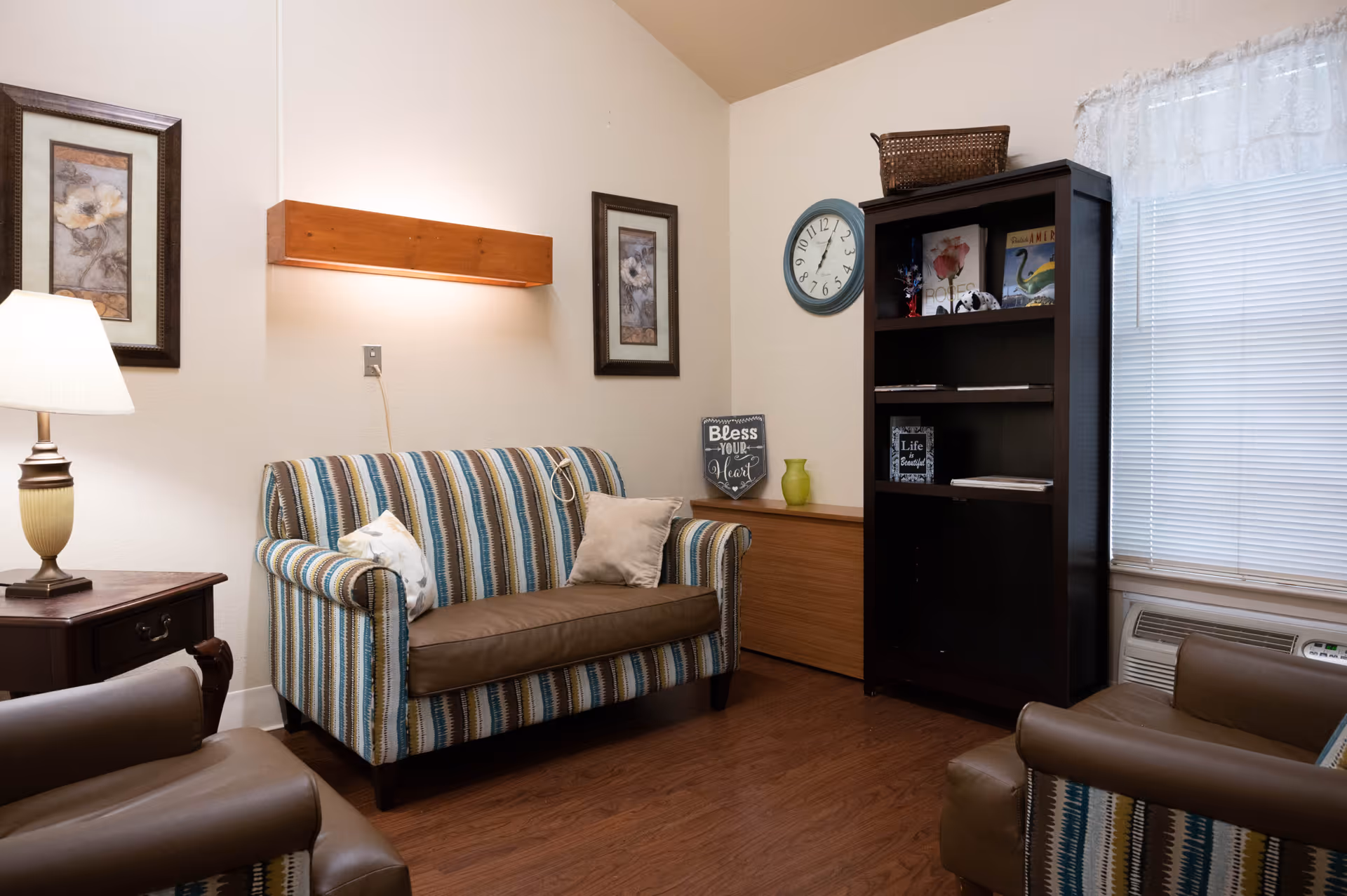 A cozy living room area with a striped loveseat and matching armchairs, a wooden side table with a lamp, framed floral artwork on the walls, a wall-mounted wooden light fixture, a black bookshelf with decorative items and books, a round wall clock, and a window with blinds and a lace valance.