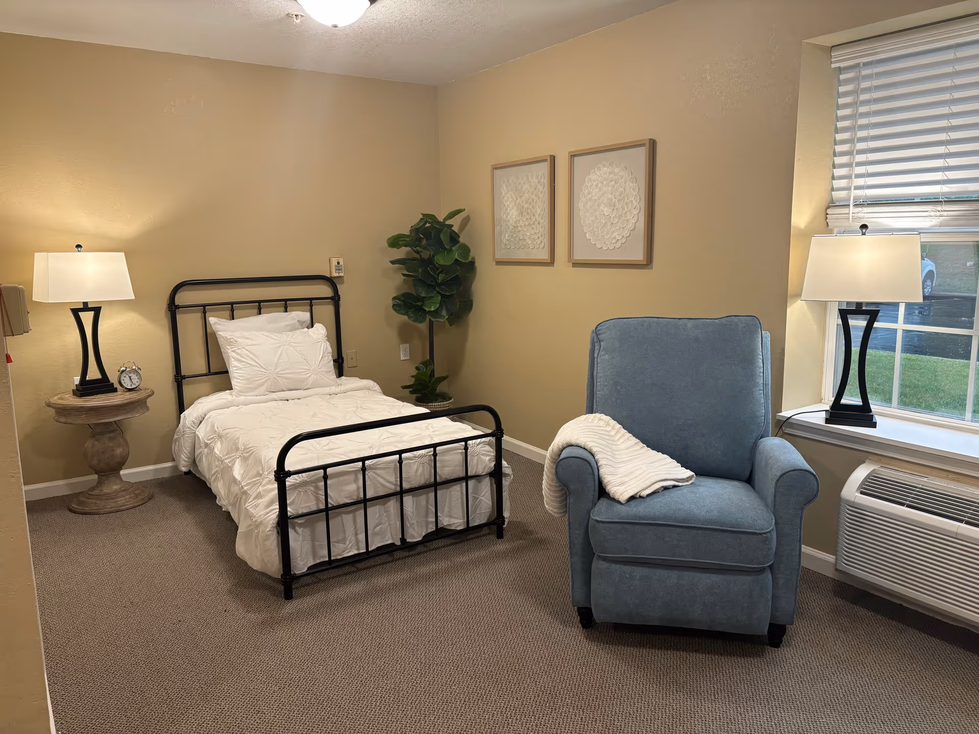 A cozy bedroom with a single black metal bed frame with white bedding, a round wooden side table with a lamp and alarm clock, a blue upholstered armchair with a white throw blanket, two framed artworks on the beige wall, a window with blinds, and a wall-mounted air conditioning unit below the window.
