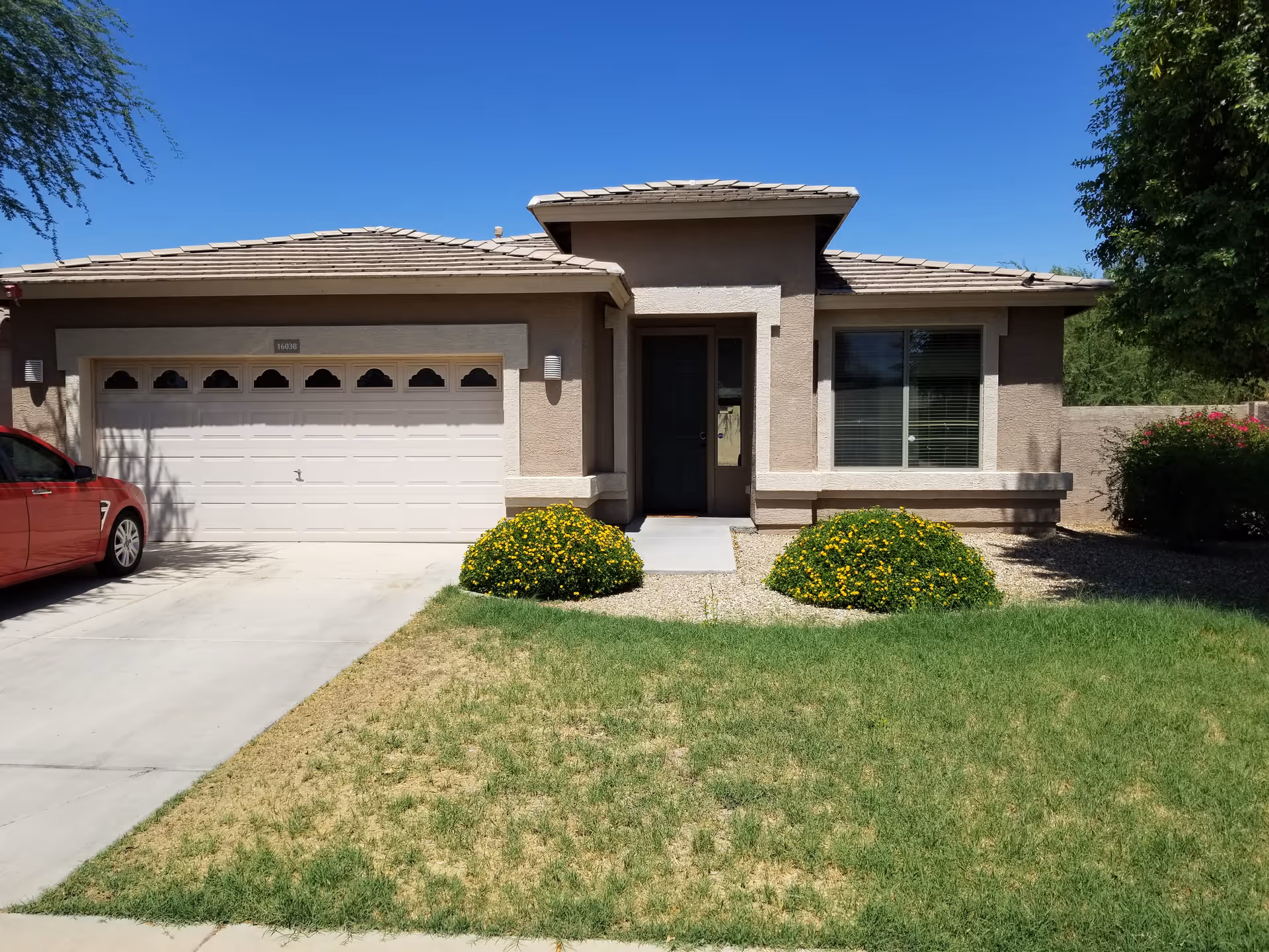 Front exterior view of a single-story house with a beige facade, a two-car garage, a dark front door, two bushes with yellow flowers, a green lawn, and a red car parked on the driveway under a clear blue sky.