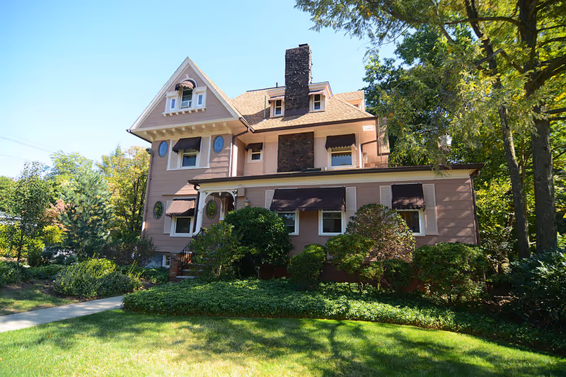 Exterior view of a large, multi-story house with beige siding, brown window awnings, and a prominent brick chimney. The house is surrounded by green bushes, trees, and a well-maintained lawn under a clear blue sky.
