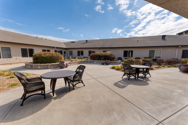 Outdoor courtyard area at Cedarhurst Senior Living of Springfield, MO with round tables and wicker chairs on a concrete patio, surrounded by low bushes and a one-story building under a partly cloudy sky.