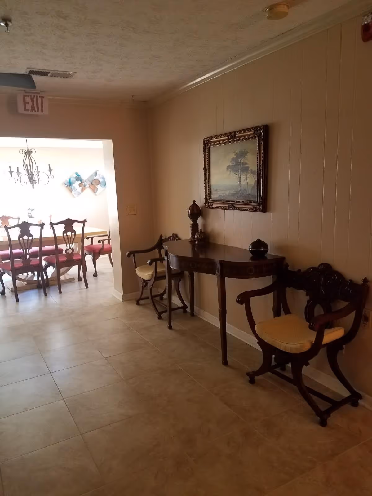 Interior hallway with beige tiled floor and light brown walls featuring a wooden table with decorative items and two wooden chairs with cushions. A framed painting hangs above the table. In the background, there is a dining area with a wooden dining table, chairs with red cushions, a chandelier, and wall decorations. An exit sign is visible on the ceiling near the hallway entrance.