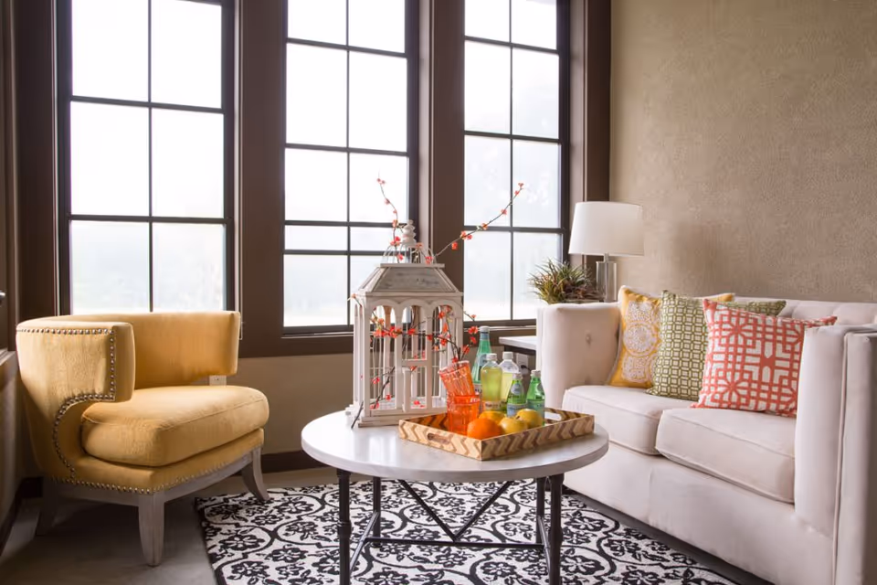 Sunlit living room corner with a beige sofa, yellow armchair, and a round coffee table holding drinks and fruit in front of large paned windows.
