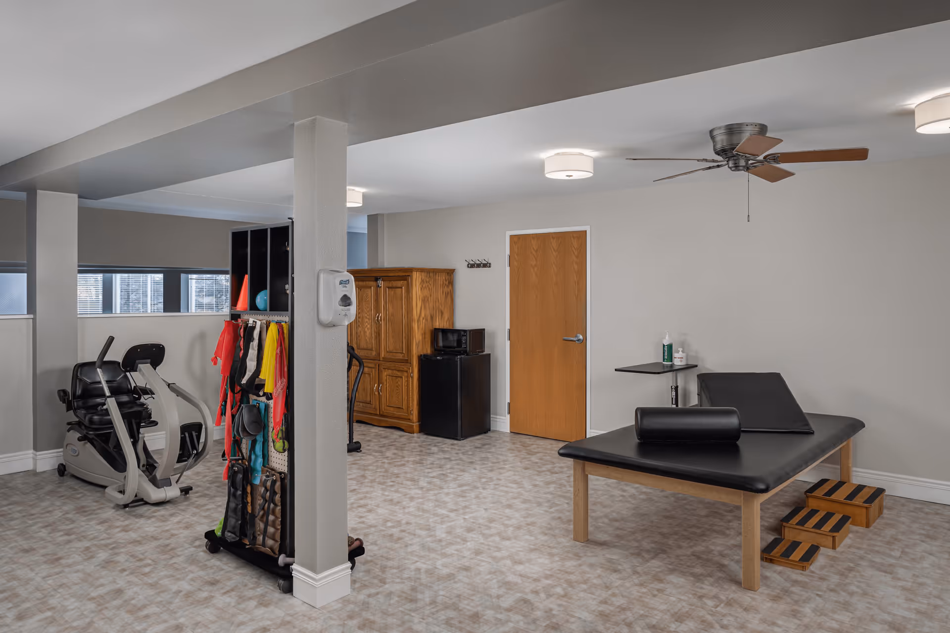 A spacious room in a senior living facility with exercise equipment including a recumbent bike, a rack holding various exercise bands and accessories, a wooden cabinet, a small refrigerator, and a black padded therapy or massage table with a bolster and incline cushion. The room has beige walls, a ceiling fan, and overhead lights.