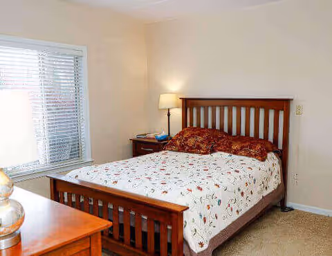 A bedroom with a wooden bed frame and a bed covered with a white bedspread featuring a floral pattern. There are two matching pillows on the bed. A wooden nightstand with a lamp and some items is next to the bed. A window with white blinds is on the left wall, and a wooden dresser is partially visible in the foreground.
