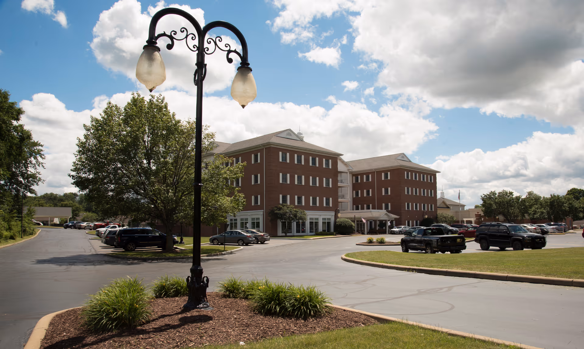 Exterior view of a multi-story brick building with several windows, surrounded by a parking lot with cars and green landscaping. A decorative black street lamp with two lights is in the foreground, and the sky is partly cloudy.