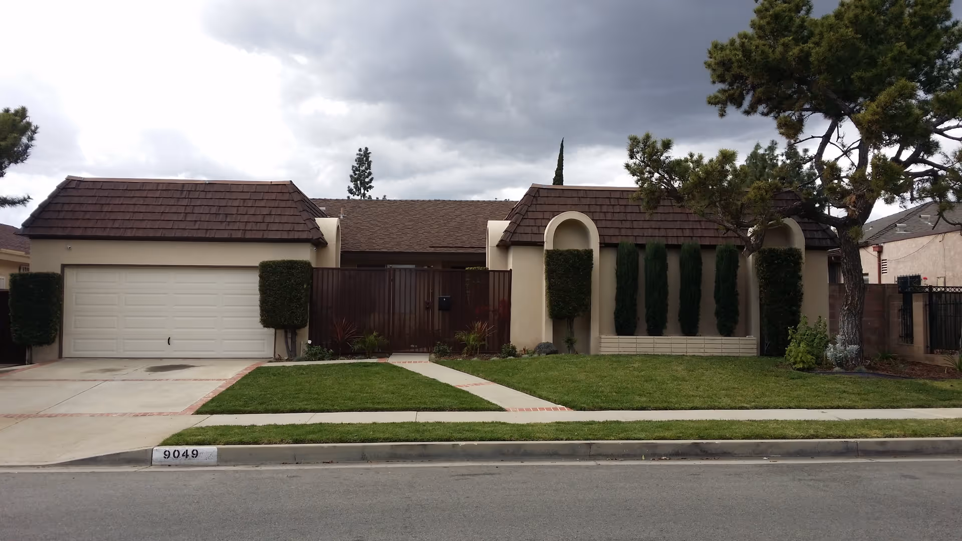 Front view of a single-story house with an attached two-car garage, gated entrance, trimmed hedges, and a lawn under a cloudy sky.