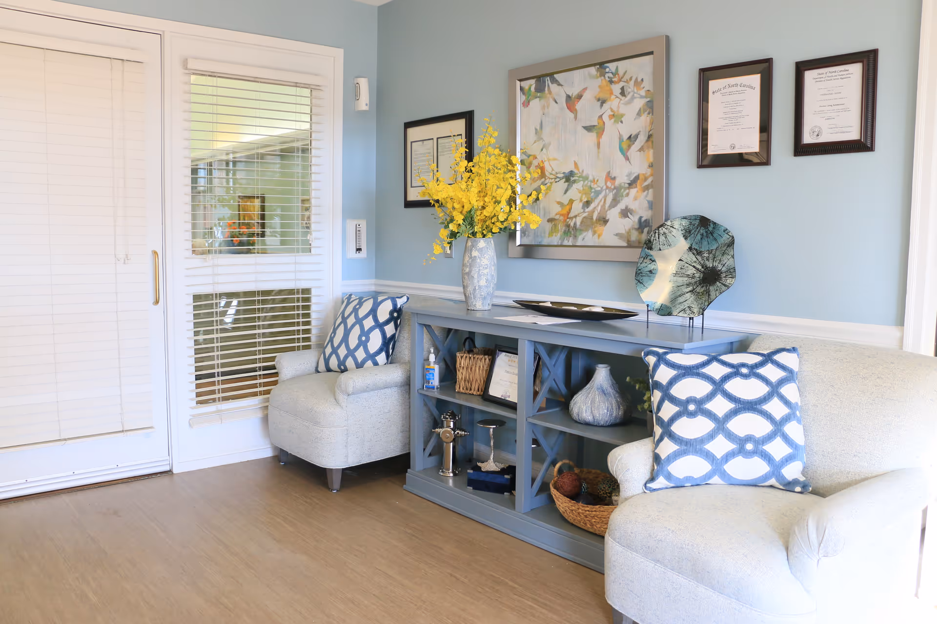A cozy sitting area in Preston House featuring two light gray armchairs with blue and white patterned pillows. Between the chairs is a blue wooden console table decorated with a vase of yellow flowers, framed artwork, decorative plates, and various small items. The walls are painted light blue and adorned with framed certificates and a colorful bird-themed painting. A glass door with white blinds is visible on the left side.