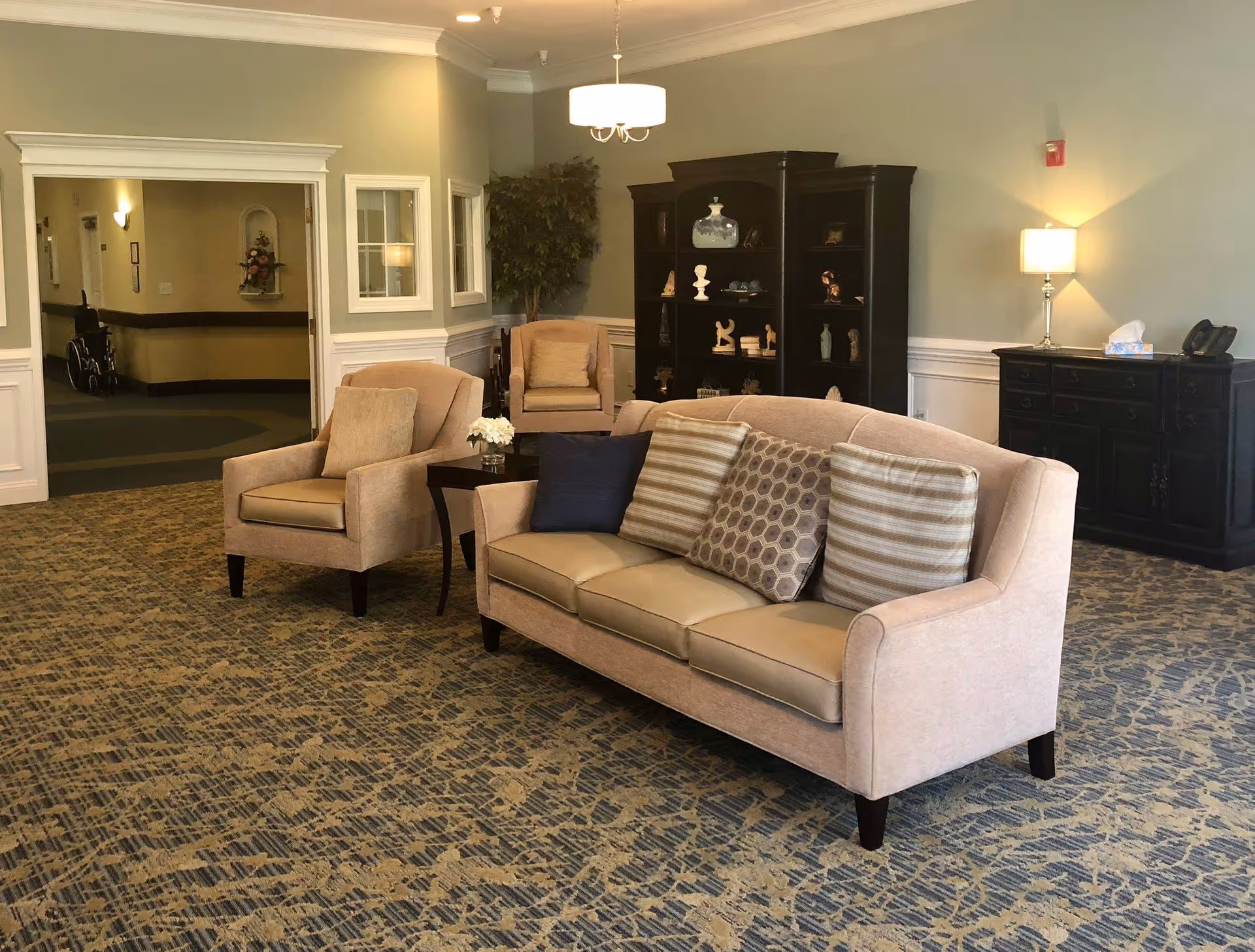 A cozy living room area in Roxboro Assisted Living featuring a beige sofa with four decorative pillows, a matching armchair, a dark wooden side table with a small flower arrangement, a large dark wooden cabinet with decorative items, a black sideboard with a lamp and tissue box, and a patterned carpet. The walls are light-colored with white trim, and there is a hallway visible in the background with a wheelchair parked.