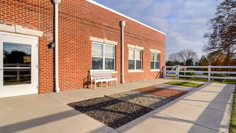 Brick exterior of a nursing facility showing a concrete patio with a white bench, windows, and a white fence under a partly cloudy sky.