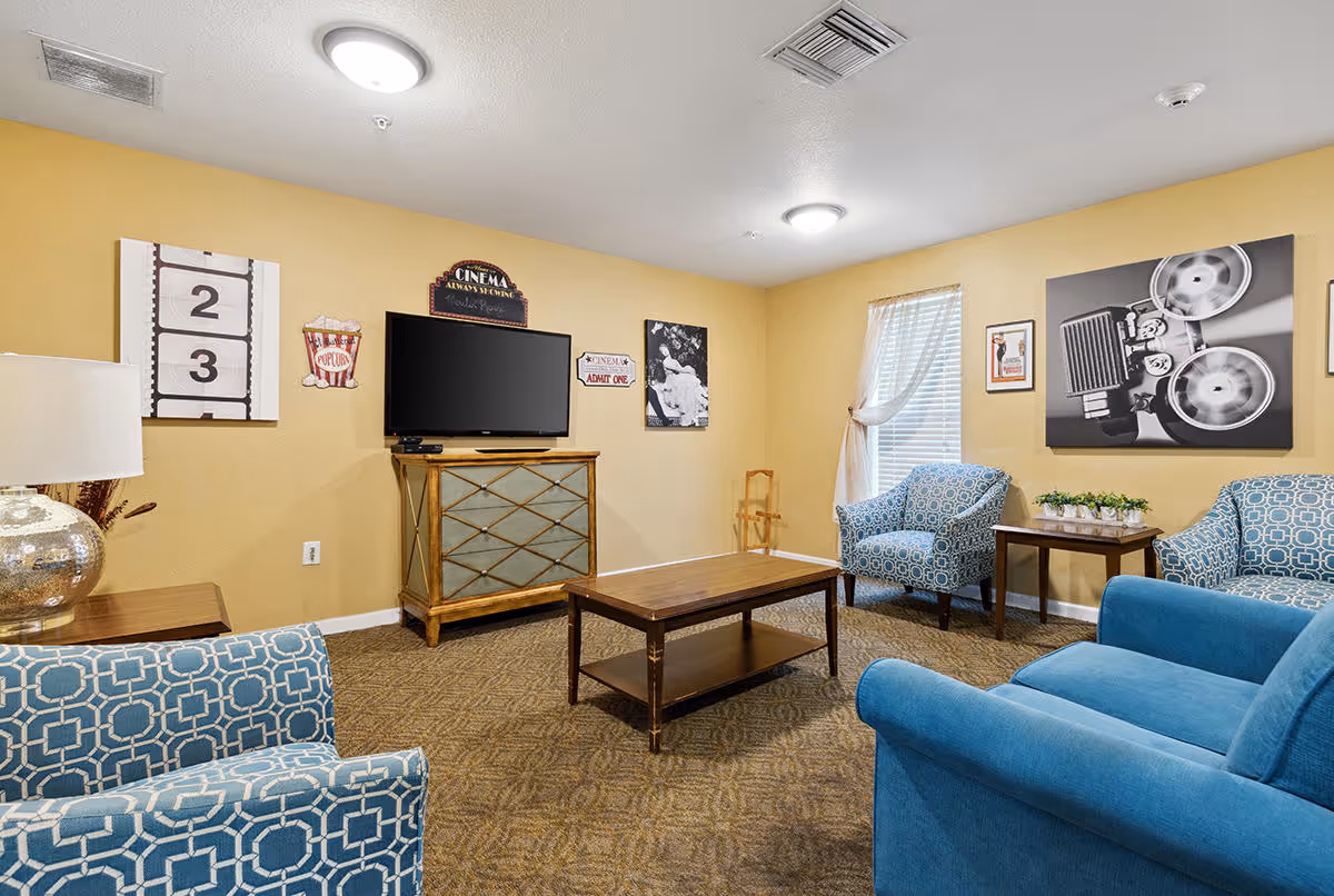 A cozy living room with yellow walls, featuring a flat-screen TV on a wooden cabinet, a wooden coffee table, and four upholstered chairs in blue and patterned fabric. The walls are decorated with vintage cinema-themed artwork and signs. A window with sheer curtains allows natural light into the room.